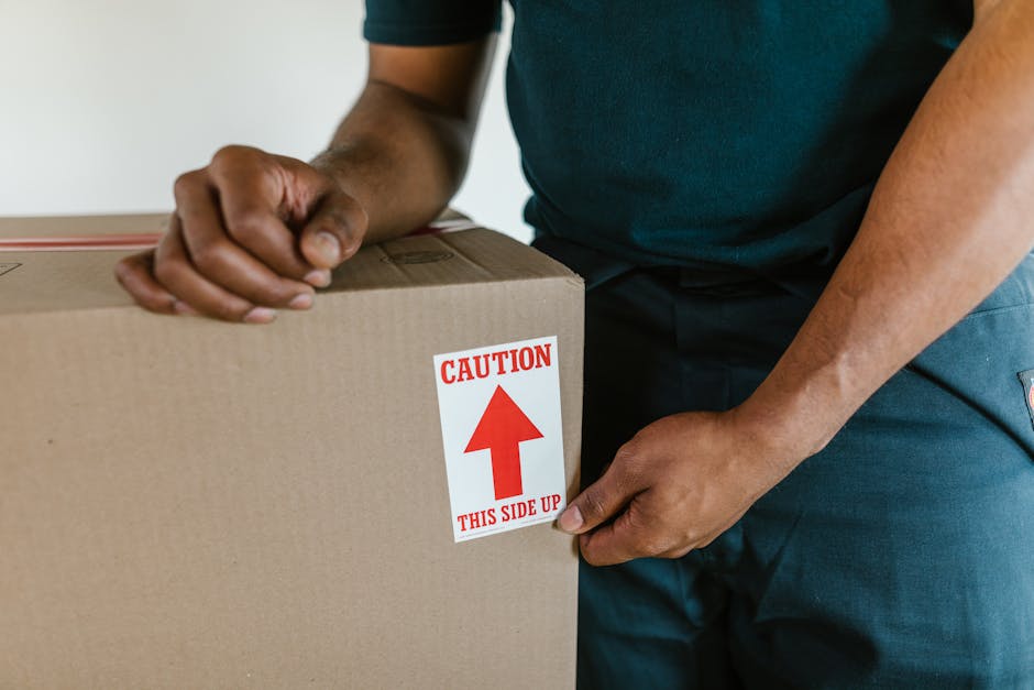 A young woman with long brown hair, dressed in a white tank top and beige pants, is sitting cross-legged on a wooden floor surrounded by numerous cardboard moving boxes of varying sizes, some sealed w
